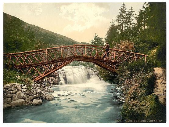The image depicts a picturesque rural scene. At the center, there is a rustic wooden bridge with metal railings, spanning across a river. A waterfall cascades in the background, adding to the natural beauty of the setting.

A wooden walkway leads up to the bridge from the viewer's perspective, suggesting an inviting path for visitors to enjoy the scenery. The bridge is set amidst a lush landscape of trees and foliage, contributing to a tranquil atmosphere.

The river flowing beneath the bridge appears calm and serene, with small rapids visible near its base, possibly indicating the presence of fish or other aquatic life. A few rocks can also be seen on either side of the waterfall.

Above the bridge, wooden supports provide stability, reflecting the robust construction of the era when this photo was likely taken. The style and coloration suggest that the image could date back to the late 19th or early 20th century. The overall composition of the photograph conveys a sense of peacefulness and timeless beauty that is characteristic of natural landscapes.