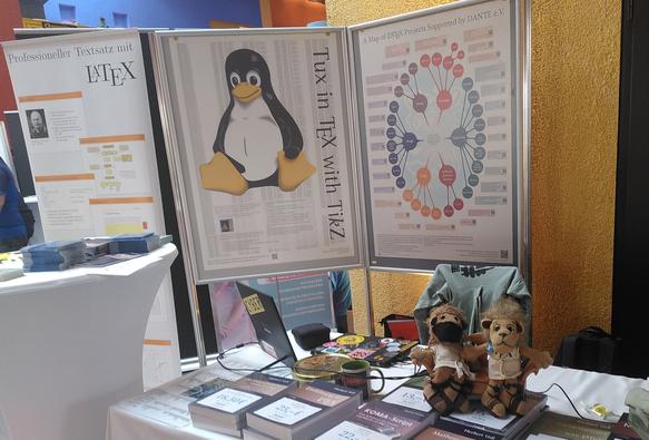 Foto des Dante Standes auf den Chemnitzer Linuxtagen 2025. im Hintergrund ein Poster „Professioneller Textsatz mit LaTeX", „Tux in TikZ“ und „A map of (La)TeX Projects supported by DANTE e.V. 

Im Vordergrund ein Tisch mit Büchern der DANTE Edition, Aufklebern, zwei Stofftier TeX Löwen und ein Laptop.
