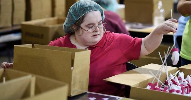 photo of a white disabled woman in a sheltered workshop, wearing a hairnet, surrounded by boxes, holding onto a spray bottle top