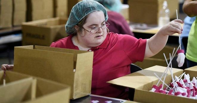 photo of a white disabled woman in a sheltered workshop, wearing a hairnet, surrounded by boxes, holding onto a spray bottle top