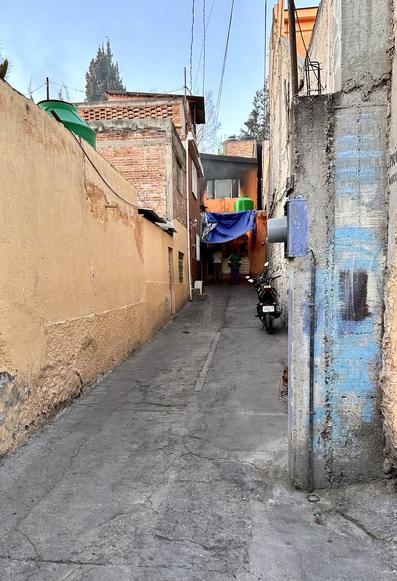 Narrow alleyway with concrete pavement flanked by weathered walls of adjacent buildings. There's a parked motorcycle, hanging laundry, and electrical wiring overhead. A person is partly visible in the distance.