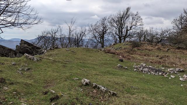 Green grass on flat part of the rocky hill ending with cliff. Some leafless trees and hills in the background.