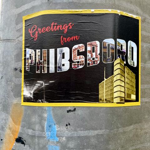 A sticker with the text "Greetings from PHIBSBORO" featuring pictures of buildings and scenery within the letters, on a black background with yellow edging, on a metal pole with spray paint splatters.