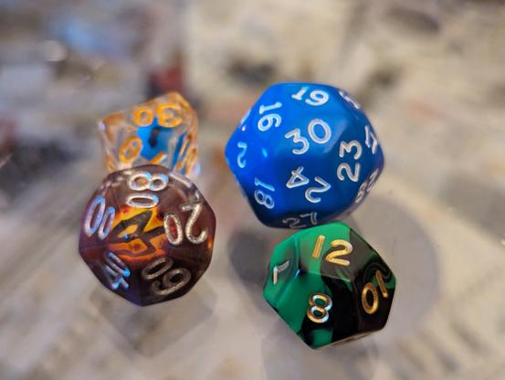 Four dice on a glass tabletop. A ten side clear die with a blue shape inside it. A second ten sided smoked dice with an eye of Sauron effect flaming eye embedded. In foreground a green and black swirled twelve sided dice with gold numbers. Dominating those is a large blue 30 sided dice with white numbers