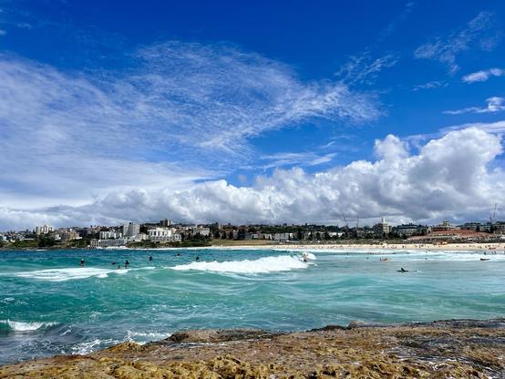 View across Bondi Beach from the north, perspective standing on the rock pools. The water is aqua blue, a large wave breaks ridden by a surfer. Others are paddling out. 
In the background the expanse of sand wraps around dotted with buildings and a long, dramatic cloud hugs the horizon