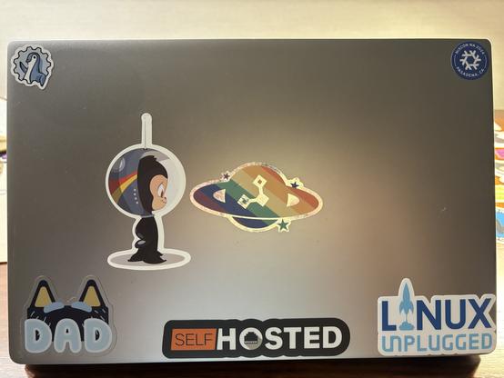 Photo of the lid of my laptop showing stickers for: SCaLE, NixCon North America, Self Hosted, Linux Unplugged, a “dad” sticker themed from Bluey, an astronaut like sticker from GitHub, and a rainbow planet sticker I got from Puppet.