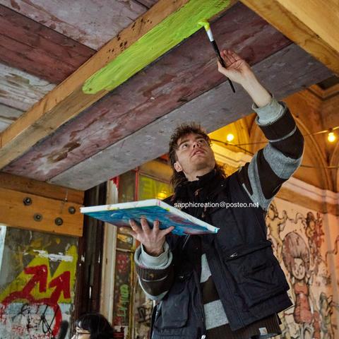 Eine Person hat Farbe auf eine umgedrehte Leinwand gefüllt und pinselt den Balken über ihr gelb an. Im Hintergrund sind ältere Wandgestaltungen zu erkennen, unter anderem ein ⚣.