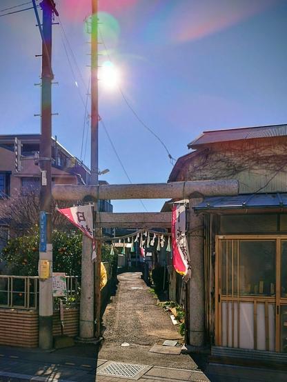 The path to the actual shrine is a narrow footpath between houses, mostly residential buildings. This photo was taken from the street the path leads off of. The stone gate is wider at the top than there is space for the path. It's windy and a lot of the colorful banners that are hung at the entrance and along the path are going in all directions. The shot is backlit and the sun is peeking out behind an electricity pole by the street causing a lense flare.