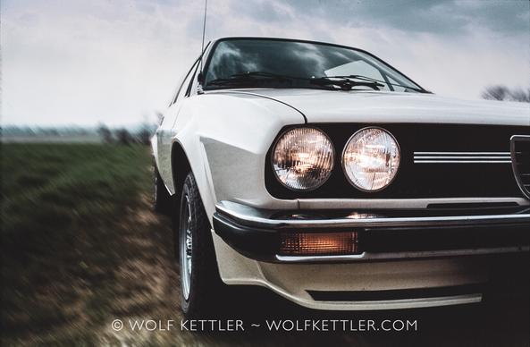 Photograph shows partial front and side of an Italian 1980s sports car from a low viewpoint. The headlights are on.