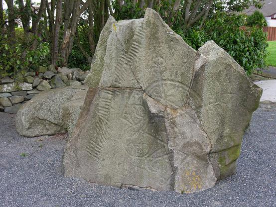 The Brandsbutt symbol stone in Inverurie. The image shows a large irregularly-shaped stone surrounded by a flat gravel surface and backed by a low stone wall with bushes. The face of the stone has a series of Pictish symbols carved into it and a nearly vertical edge in the centre of the frame has a column of short horizontal markings wrapping round the two sides of a corner.