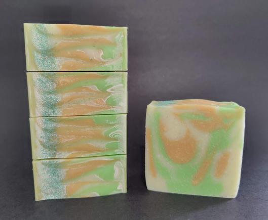 On the Left are 4 bars of cream, green and gold soaps stacked on their side to show the top, which has a sprinkling of green jojoba pearls. with a single bar on the right.