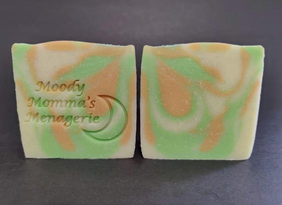 Two bars of cream, green and gold soaps sitting side by side. The bar on the left is stamped with "Moody Momma's Menagerie" and a crescent moon