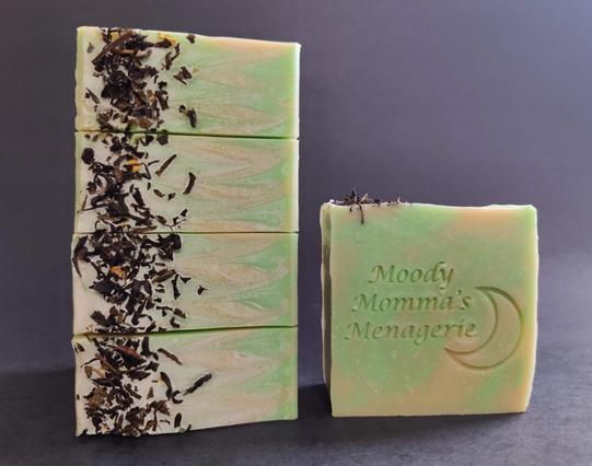 On the Left are 4 bars of green and gold soaps stacked on their side to show the top, which has a sprinkling of green tea leaves. The single bar on the right is stamped with "Moody Momma's Menagerie" and a crescent moon