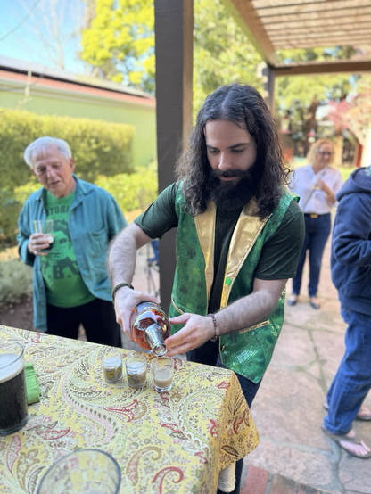 A young man with long hair, beard and mustache and green vest pours Jameson whiskey into glasses containing Irish Cream liqeuer. Later they will be dropped into Irish stout and slammed to the head.