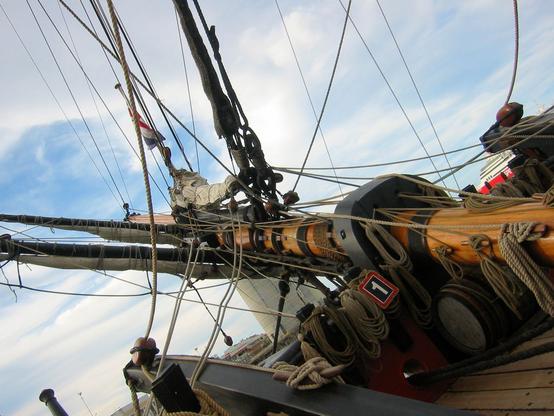 The bowsprit of a large ship (that's the mast the sticks horizontally out the front).  There are rope and cables running from it at in all directions, and some more coiled and hanging near the base. Two yards are sticking off to the side, with sails furled tightly against them.  In the bottom right corner are some boards of the deck, rather than being sensible at the bottom of the photo.
In the background can be seen part of a large white and red ship (Spirit of Tasmania ferry)  and a large white silo (I think it holds coal or something like that).
The sky is mostly white, with fluffy clouds, but there some patches are blue.