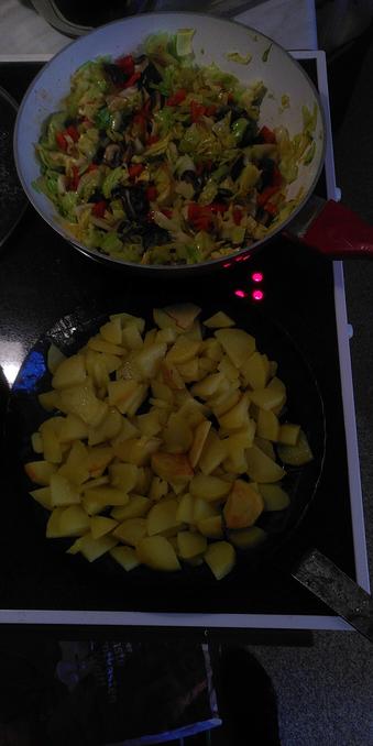 Zwei Pfannen von oben auf dem Herd gesehen. Eine mit Bratkartoffeln, die andere mit Spitzkohl, Champignons und Paprika.