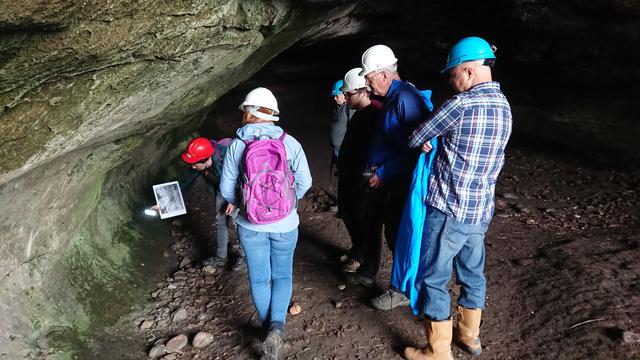 A group of people in hard hats standing in a cave, with one bending down, holding a picture and pointing a light at the wall.