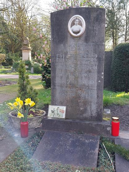 Foto vom Albert Richters Grab (nachdem ich die Blumen und Kerze hingestellt habe).  
Ein schlichtes Einzelgrab mit einem dauerhaft befestigtem Portraitfoto von ihm im Radtrikot. Der Grabstein besteht aus Beton, die Gravur lautet: Albert Richter Rennfahrer *14.10.1912 +2.1.1940 „Wer dich gekannt / vergisst dich nie“.
An den Grabstein lehnt eine Postkarte von Albert Richter auf dem Rennrad mit der Aufschrift "With respects from Canada":  Auf dem Grabstein liegen einige kleine Kieselsteine. Links vor dem Grabstein steht ein kleines Bäumchen in einer grauen Schale. 

Jetzt steht vor der Schale mit dem Bäumchen ein Strauß gelber Narzissen in einem roten Gefäß. Rechts vom Grabstein steht ein brennendes rotes Grablicht mit Metalldeckel.