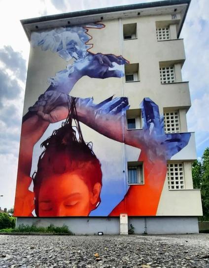 Streetartwall. On the exterior of a three-story modern residential building is sprayed/painted mural of a portrait of a woman. The background is a light beige house color and the mural is designed in shades of red, blue and white. You can see half the face of a dark-haired woman designed in red, raising her arms in the air as if she were immersed in water. The arms are surrounded by blue water, which turns into crystals on the skin and grows there.