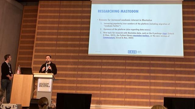 Two researchers presenting on a large stage with a slide displayed entitled: researching Mastodon