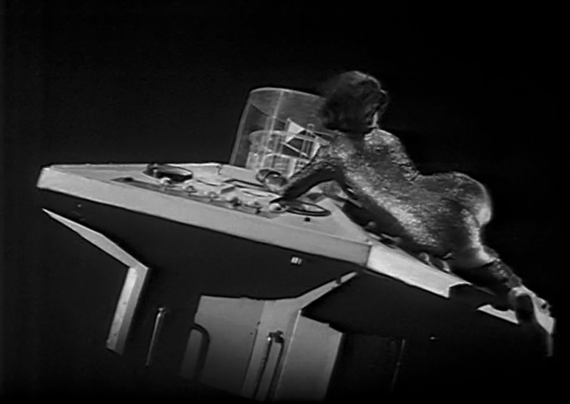 The centre console of the TARDIS is floating in space at an angle (dipped to the bottom right corner). 
Zoe, wearing a figure hugging glittery catsuit with her back towards the camera, is holding onto the centre column for dear life.