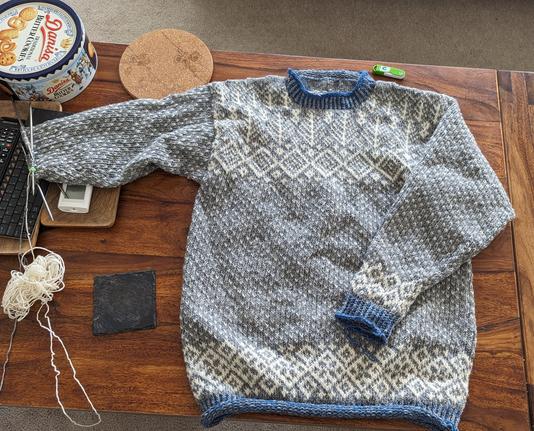 A knitted jumper lies on a wooden table, one sleeve is incomplete and still has double pointed needles attached.  It is a grey and stranded pattern