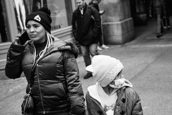 The image shows a black and white street scene featuring two main subjects: a woman and a young child. The woman is on the left side of the frame, wearing a winter hat and a puff jacket. She appears to be on the phone, holding a mobile device to her ear with a focused or concerned expression on her face. The young child on the right is wearing a light-colored hat and a patterned scarf or mask covering their mouth and nose, along with a coat. In the background, other individuals can be seen walking down the street, suggesting an urban environment. The overall image captures a candid, everyday moment on a city street, where the individuals appear to be going about their daily routines.