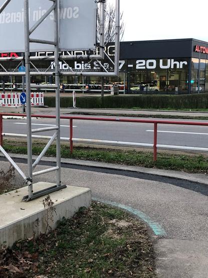 Zu sehen ist ein Radweg mit ca. 1,5 m Breite und mit einer 90° Kurve. In der rechten Hälfte des Radwegs verläuft ein ca. 40 cm breiter, noch nicht endgültig mit der Deckschicht verschlossener Graben, sodass dort eine scharfe Kante von ca. 5 cm ist, damit ist die rechte Hälfte des Radwegs nicht nutzbar.
An der Innenseite der Kurve ist ein Betonsockel, auf dem das Gestell für eine große Hinweistafel der angrenzenden Baustelle steht.
In ca. 1,7 m Höhe ist die Ecke der Hinweistafel direkt an der Kante des Radwegs.