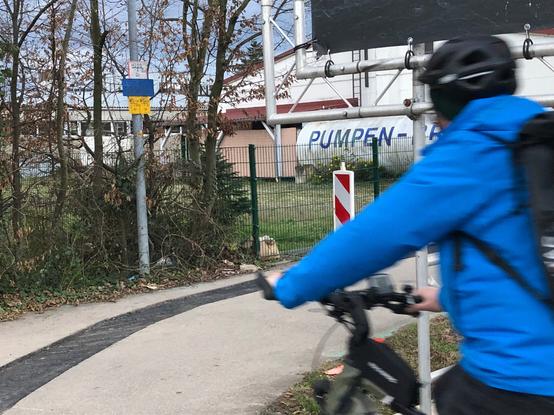 Hier ist wieder der Radweg mit der Kurve und dem nicht vollständig geschlossenen Graben zu sehen, diesmal aus der anderen Richtung. Ein Radfahrer in blauer Jacke, mit schwarzem Rucksack und schwarzem Helm ist kurz davor, der Rechtskurve im Radweg zu folgen. Gut zu sehen ist, dass die massive Ecke (aus Rundrohren) der Hinweistafel genau in Kopfhöhe des Radfahrers ist und dass die Ecke gefährlich nahe am Radweg ist.