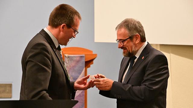 Eine Silbermedaille bildet die Mitte der Szene: Der Herr in der linken Bildhälfte, Mario Fischer (schwarzes Jackett, Brille, Kurzhaarschnitt), überreicht dem Herrn in der rechten Bildhälfte, Bischof Harald Rückert (schwarzes Jackett, Bart, Brille), die Medaille.