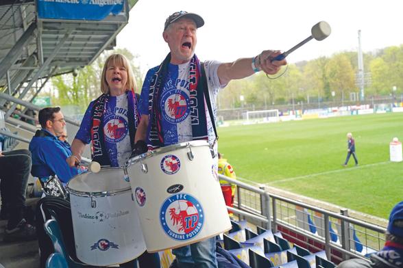 Die Fantribüne des Stadions von Turbine Potsdam, rechts im Hintergrund der Rasen, auf dem verschwommen eine Person läuft. Links im Vordergrund eine Frau und ein Mann in Turbine-Kluft, beide tragen Schals um den Hals und große Trommeln vorm Körper. Es ist erkennbar, dass sie singen oder anfeuern. Die Frau schlägt ihre Trommel, der Mann hält den Klöppel ausgestreckt vor sich.