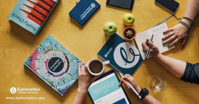 Photo looking down on a table with a collection of data books, Epimorphics notepad, coffee mug, apples, people’s hands taking notes and an iPad with our website plus the URL: www.epimorphics.com