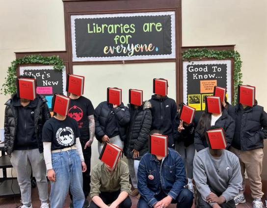 3 warm up organizers, a teacher, and 9 students standing in a library under a sign saying, ‘Libraries are for everyone.’