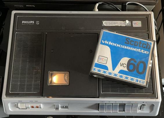 Un magnétoscope VCR Philips N1481 de 1972 en cours de lecture d'une cassette.
Il y a le boitier d'une cassette Scotch VC60 posé sur le magnétoscope.
On voit de la lumière sur la fenêtre de la trappe à cassette qui permet de voir combien de bande est déjà passé.