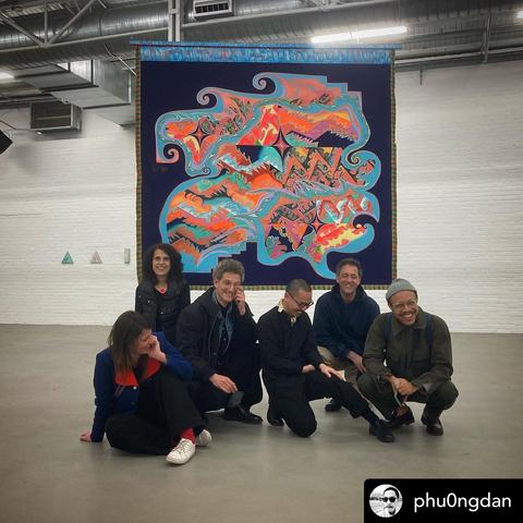 5 artists /  curators / 2 women 4 men,  in Kunsthalle Münster May 2023 exhibition ton not. not ton. grey floor, white walls, silver pipes at the ceiling, a blue / orange / yellow / cyan colored flag painting of Lisa Alvarado behind the people