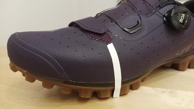 Eine Nahaufnahme von einem lila-farbenen Mountainbike- und Gravel-Schuh mit schwarzem BOA-Verschlusssystem. An der Schuhaußenseite ist ein weißer Klebestreifen angebracht.