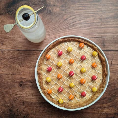 A photo of a pie and a cocktail on a dark wooden background, taken from above. The pie is a frangipane-filled sweet shortcrust. It is decorated with wee balls of marzipan in yellow, orange and red colours, with a grid of of white icing over the top. The cocktail is a gingery-yellow coloured drink in a highball glass filled with ice, with a garnish of a hollowed-out lemon peel surrounding a cherry on a cocktail pick (vaguely looking like Saturn and its rings).