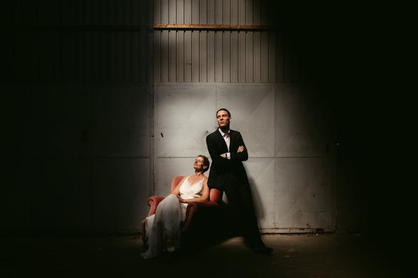 This image conveys a dramatic and artistic portrayal of a wedding couple in Vienna. The setting appears to be an industrial or warehouse space, with the texture of raw concrete on the floor and walls. There's a stark contrast of light and shadow, with a beam of light illuminating the couple while the surroundings are shrouded in darkness.

The bride is seated on a vintage pink armchair. She's dressed in a graceful white gown that drapes elegantly over the chair and onto the floor. Her posture is relaxed, her face tilted upwards towards the light, eyes closed, with a serene expression that suggests a moment of peaceful introspection.

The groom stands next to her, exuding confidence and contemplation. He's dressed in a classic black suit and white shirt, his arms crossed over his chest. His gaze is directed out of frame, possibly reflecting on the significance of the day.

The composition is thoughtful, with the placement of the couple off-center in the frame creating an intriguing visual tension. The minimalist setting focuses all attention on the couple, their expressions, and their elegant wedding attire, making the image a powerful statement of their bond and the ambiance of their special day.