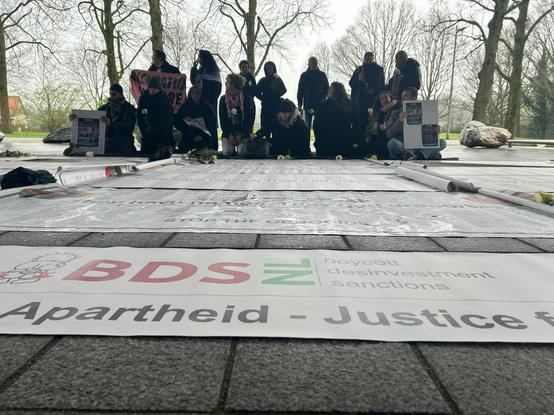 Op de grond liggen banners, de voorste is een bannering van BDS. De woorden apartheid  en justitie zijn te lezen. Daarachter liggen banners met namen van vermoorde Palestijnse burgers. In de achtergrond  zit en staat een groepje mensen in zwarte rouwkleding.