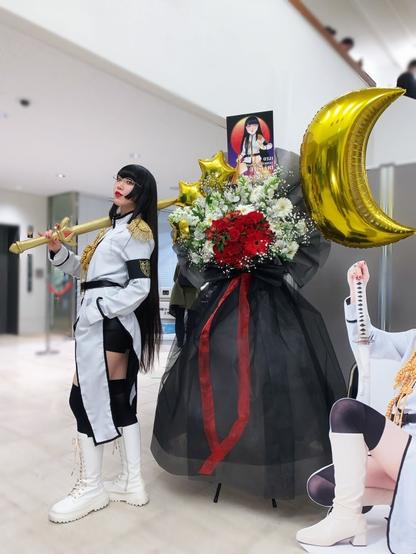 Himari dressed as Persephone stands next to a large flower stand.