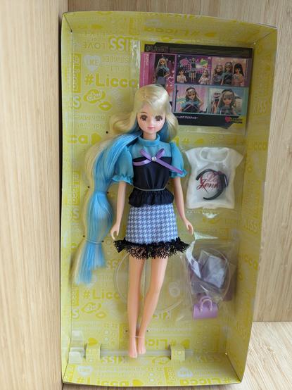 A photo of Hashtag Jenny still attached to her cardboard box insert. She is attached to the cardboard with plastic tags that loop around her neck, waist, and feet. Her accessories and shoes are in plastic bags that are taped to the cardboard backing.
