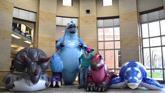 An anthropomorphic sabertooth in a teal shirt stands in a rotunda with four oversized mesozoic inflatables--a grey triceratops tube, a blue standing tyrannosaurus, a maroon laying raptor, and a blue pliosaur.