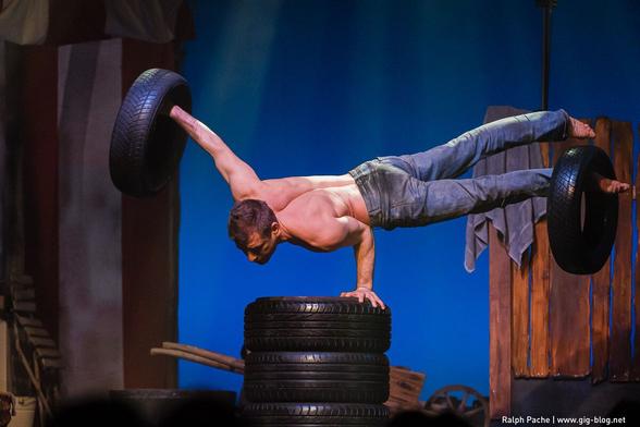 Ein Artist macht einen Ein-Arm-Stand, während er am anderen Arm und einem Bein einen Autoreifen balanciert.
