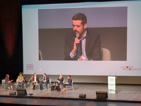 4 hommes et 2 femmes sont assis sur scène. E. Hardy a la parole et est rediffusé sur l'écran géant.