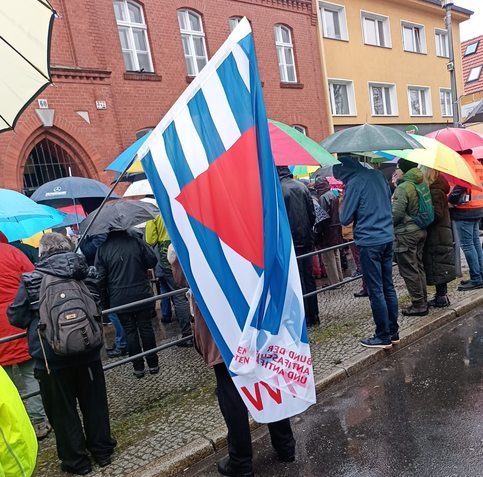 Blick auf die Dorfschule, davor stehen etliche Leute mit Regenschirmen. Eine Frau trägt die Fahne der Vereinigung der Verfolgten des Naziregimes – Bund der Antifaschistinnen und Antifaschisten - blau-weiße Streifen und ein rotes Dreieck