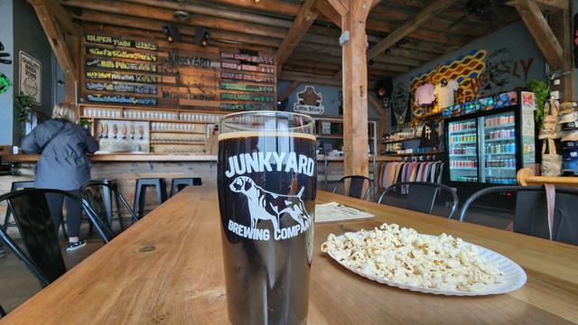 Popcorn and a pint of dark peanut butter stout in a glass with Junkyard Brewing logo set on a table with a folksie beer list on the wall. Timber framed roof and pists. A patron stands at the bar.