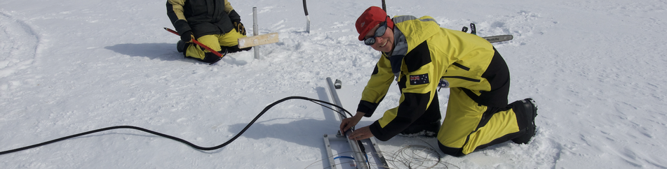 Antarctic scientist (female) working on snow, connecting solar panel cables, with other team members nearby.