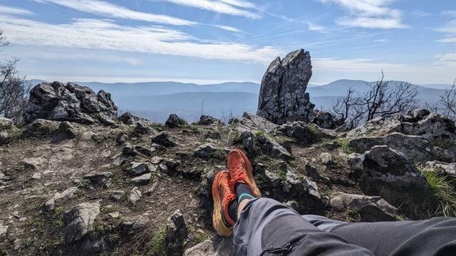 View over the edge of cliff to the distant hills. Two rocks on each side, one lower the other higher. From the right bottom corner of the photo are two legs stretched direction center in the resting position. Grey outdoor shorts, orange/yellow running shoes 😅