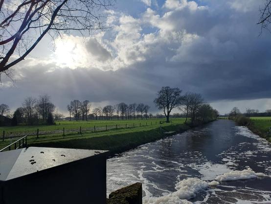 Foto van kanaal met stromend water, zon, blauwe lucht, grijze wolken en regenbuien