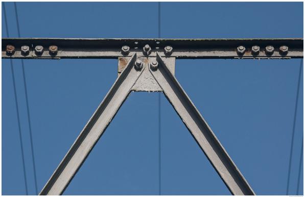 A close up colour photograph of part of an electricity pylon, which has been roughly coated in thick grey paint, showing some thirteen nuts and bolts where the structure's girders meet and are bolted together at a square plate bracket. Taken against a blue sky cloudless, in which five cables can also be seen.